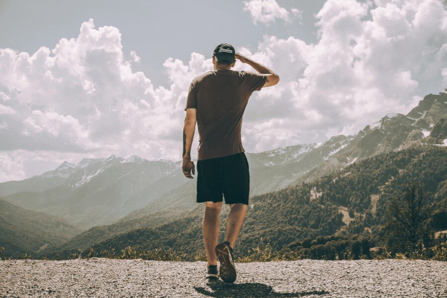 Férfi pólóban és rövidnadrágban áll háttal a fényképezőgépnek, kilátással a hegyi tájra. Foto: https://www.pexels.com/cs-cz/foto/hory-priroda-obloha-muz-2577706/