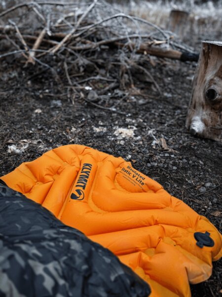 Felfújható Klymit Insulated Static V Lite matrac narancssárga színben a tűzrakóhely mellett, részben álcázó mintás hálózsákkal letakarva. Szigetelés a hideg és nedvesség ellen terepen való alváskor. Fotó: Rigad