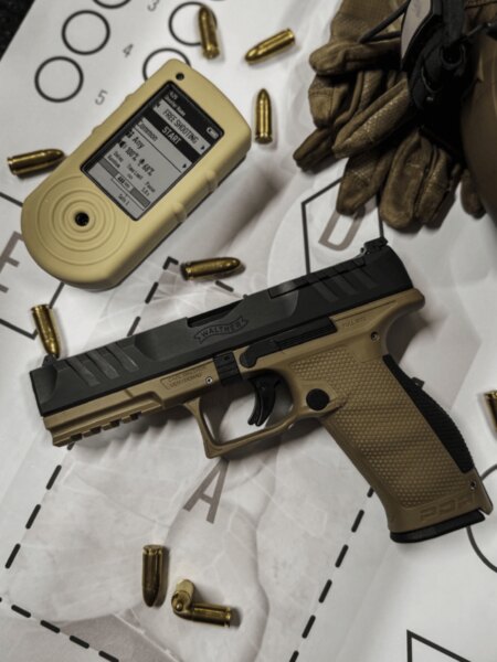 Walther PDP Full Size pisztoly lövészeti időmérővel, lőszerekkel és kesztyűkkel a céltáblán.
