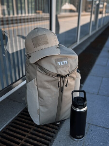 YETI Crossroads 27 l zöld hátizsák kalappal, napszemüveggel és YETI Rambler fekete edénnyel.
